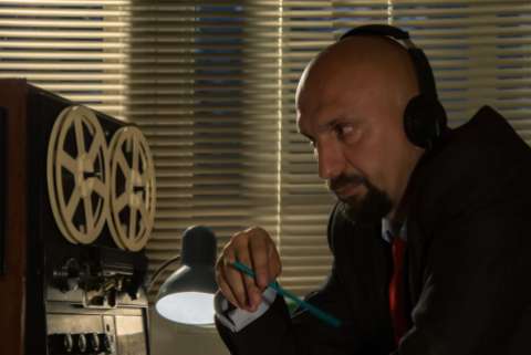 Bald man with a goatee wearing a suit and listening to recorded conversations on a reel-to-reel tape recorded and holding a pencil in a dark room