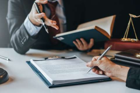 Two people conduct legal business from opposite sides of a desk, only their hands are visible, one signs a document while the other holds a book and gestures