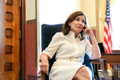 New York Governor Kathy Hochul seated at a desk. There is a flag in the background. She looks nonplussed.