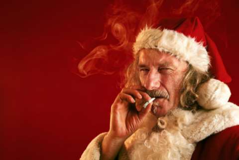 A seedy looking man in a Santa Claus outfit smokes a cigarette and angrily squints