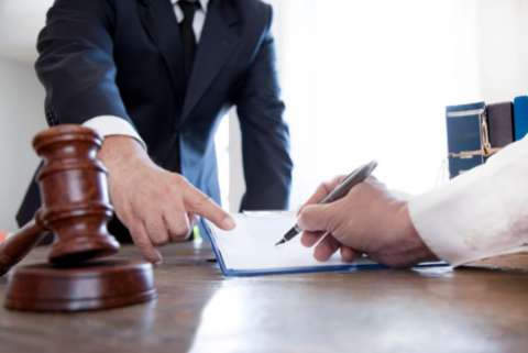 A hand signs a contract while a man in a suit points at the contract, there is a gavel on the table