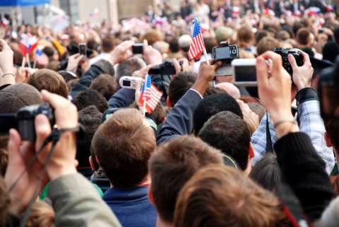 A crowd of people take photos and wave tiny American flags at a political campaign event