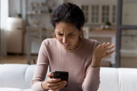 A woman seated on a couch looks at her phone, makes a disgusted face, and incredulous hand gesture