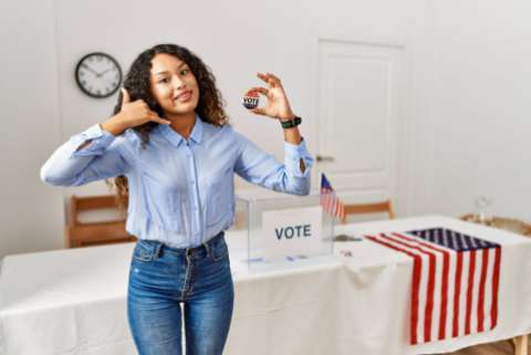 woman standing by at political campaign by voting ballot smiling doing phone gesture with hand and fingers like talking on the telephone and holding a patriotic button that says, "vote"