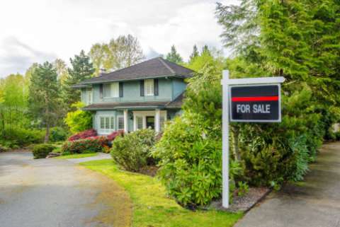 A "For Sale" sign in front of a green, two-story house surrounded by trees and bushes