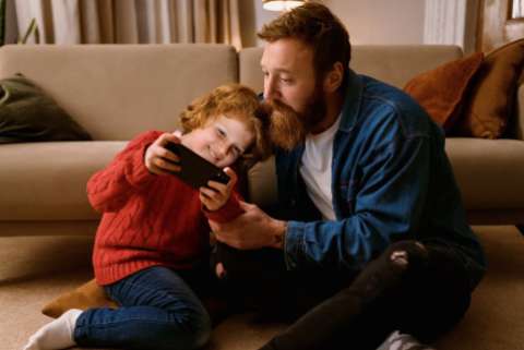 Father and son playing online game on mobile phone together while sitting on floor