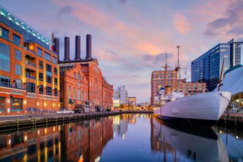A boat sits in the water in Baltimore's inner harbor at dusk, buildings new and old are visible in the background