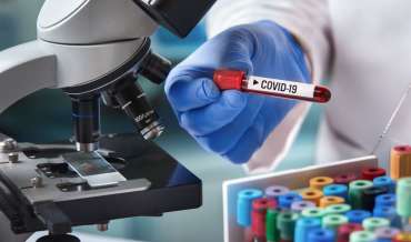 A scientist holds a vial labeled "COVID-19" in front of a microscope