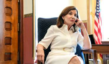 New York Governor Kathy Hochul seated at a desk. There is a flag in the background. She looks nonplussed.