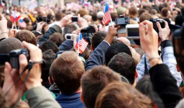 A crowd of people take photos and wave tiny American flags at a political campaign event