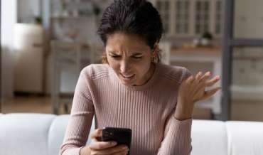 A woman seated on a couch looks at her phone, makes a disgusted face, and incredulous hand gesture