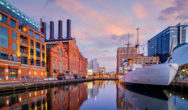 A boat sits in the water in Baltimore's inner harbor at dusk, buildings new and old are visible in the background