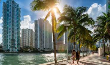 Two people walk under palm trees alongside a narrow waterway in Miami, high rises are visible on the other side of the water
